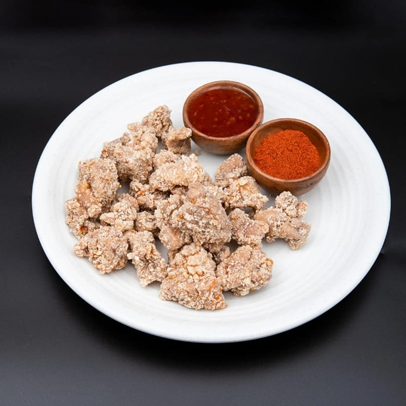 Crispy fried chicken pieces on a white plate, with two small bowls of red chili sauce and powder.